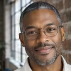 Portrait of a man with close-cropped grey-flecked hair and glasses against an exposed brick wall.
