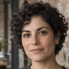 Portrait of a woman with short dark curly hair against an exposed brick wall.