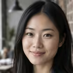 Portrait of a woman with long straight black hair in a denim shirt against an exposed brick wall.