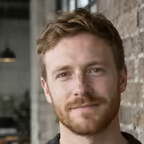 Portrait of a man with a short ginger beard in a charcoal jumper against an exposed brick wall.